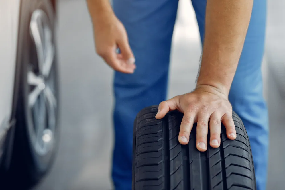Mecânico de uniforme azul com a mão apoiada sobre um pneu, ao lado de um carro